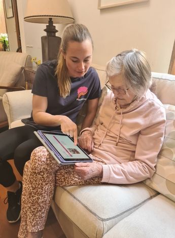 The image is of Plasticise co-founder Sarah Turner showing an older client the app on a tablet. Both women are sitting on a couch. 